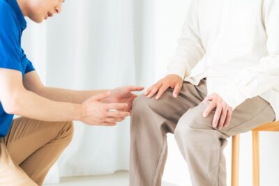 膝に手を添えて座る高齢の男性と、その膝の状態を確認するために前にしゃがみ込むスタッフ。男性は椅子に座り、スタッフは両手を差し出しながら膝の様子を観察している場面
