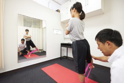 運動用ゴムバンドを足に装着し、鏡を見ながら片脚を横に上げてトレーニングする女性と、その動きを後ろからサポートするトレーナー